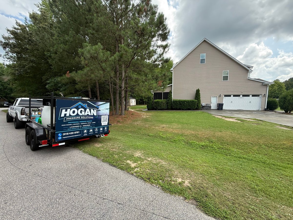 A Hogan service truck parked in the driveway of a two-story residential house with a white garage, surrounded by trees and green lawn.